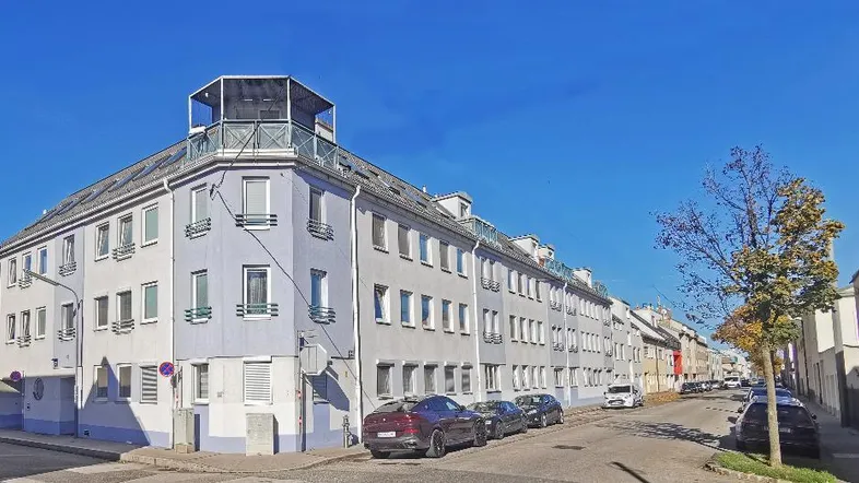 Mehrfamilienhaus mit heller Fassade, Dachterrasse und parkenden Autos in städtischer Umgebung.