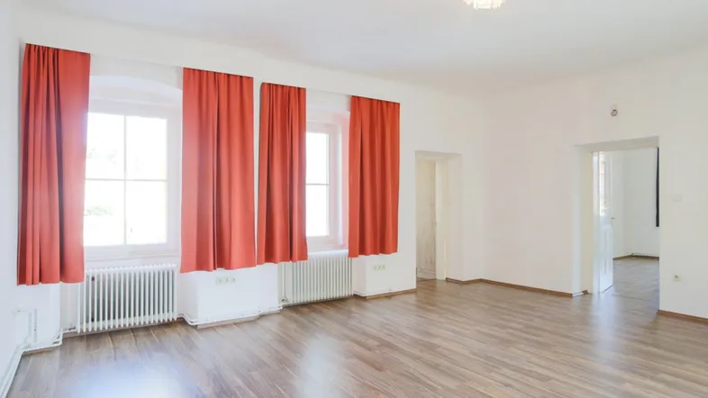 Helles Zimmer mit Holzfußboden, roten Vorhängen und Heizkörpern unter den Fenstern.