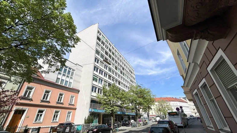 Blick auf die Straße mit einem hohen Gebäude und umliegenden Häusern unter blauem Himmel.