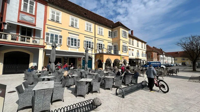 Belebter Stadtplatz mit historischen Gebäuden und Außengastronomie unter blauem Himmel.