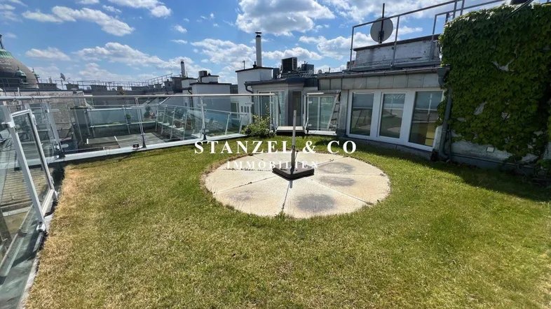 Dachterrasse mit Rasenfläche, Glasgeländer und weitem Blick über die Dächer der Stadt.