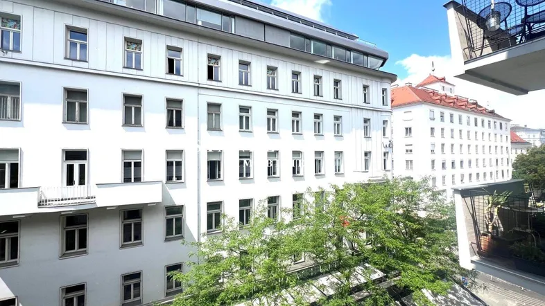 Blick auf die Stadtfassade mit einem möblierten Balkon und grünen Bäumen im Vordergrund.