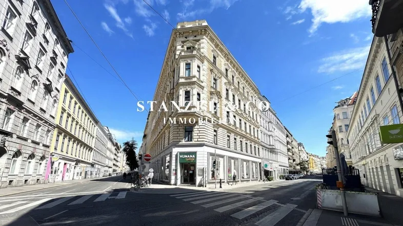 Ornates Eckgebäude an einer belebten Stadtkreuzung mit klassischer Architektur und Geschäften im Erdgeschoss.
