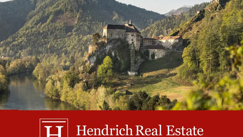 Historische Burganlage an einem Flussufer, umgeben von grünen Hügeln und Bergen.