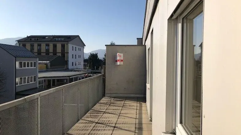 Langer Balkon mit Metallgeländer und Blick auf umliegende Gebäude und Berge.