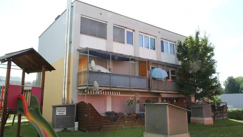 Mehrfamilienhaus mit Balkonen, Garten und Kinderspielplatz unter strahlend blauem Himmel.