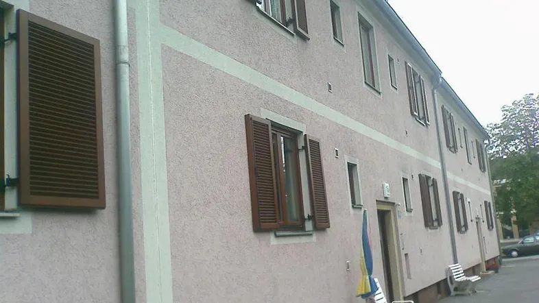 Fassade eines Wohngebäudes mit hellrosa Putz, braunen Fensterläden und einem Regenrohr.