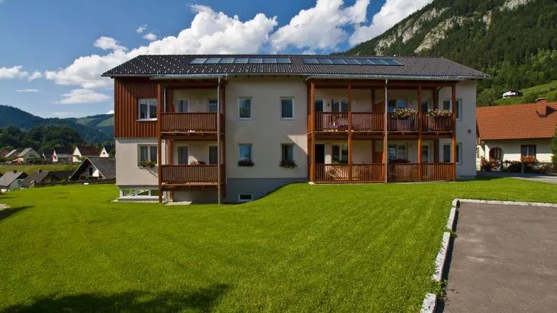 Modernes Mehrfamilienhaus mit Holzfassade, Balkonen und Photovoltaikanlage vor alpiner Bergkulisse.