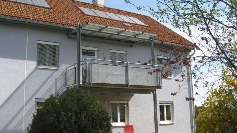 Gebäudeansicht mit Balkon, Solarpaneelen auf dem Dach und grüner Umgebung.
