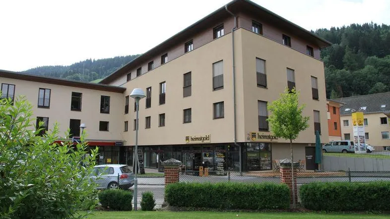 Mehrstöckiges Gebäude mit beiger Fassade, Geschäftsflächen im Erdgeschoss und Wohnungen darüber, umgeben von Grünflächen und mit Blick auf bewaldete Hügel.