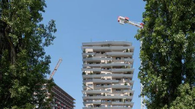 Modernes Hochhaus mit vielen Balkonen, umgeben von Bäumen und mit Baukränen im Hintergrund.