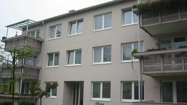 Modernes Mehrfamilienhaus mit hellbeiger Fassade, großen Fenstern und Balkonen mit Glasüberdachung.