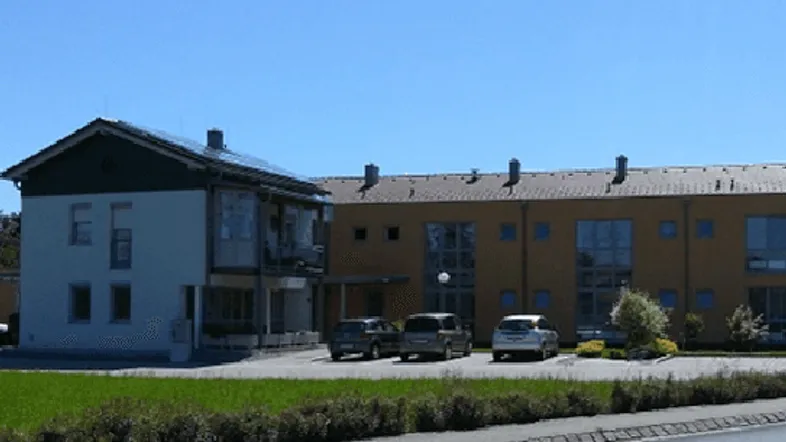 Mehrfamilienhaus mit zwei Gebäudeteilen, Photovoltaikanlage auf dem Dach und Parkplätzen davor.