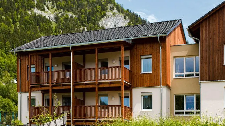 Mehrstöckiges Gebäude mit Holzfassade und Balkonen vor einer bewaldeten Bergkulisse und grüner Wiese.