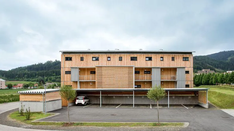 Modernes Mehrfamilienhaus mit Holzfassade, Balkonen und Carport, umgeben von grüner Landschaft.