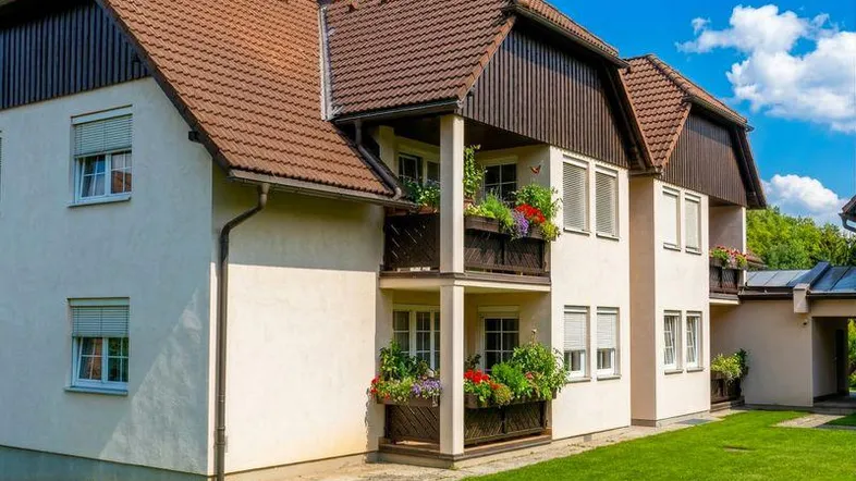 Mehrfamilienhaus mit heller Fassade, braunem Dach und blühenden Balkonen.