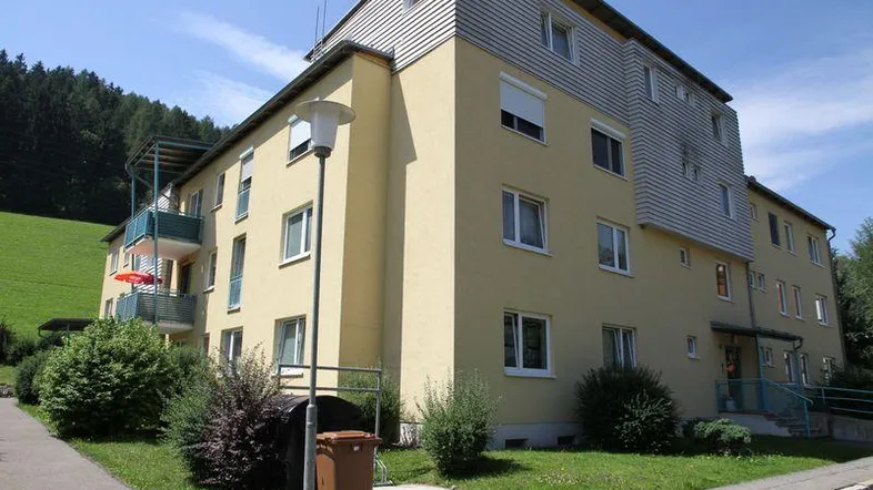 Mehrfamilienhaus mit gelber Fassade, Balkonen und grüner Umgebung unter blauem Himmel.
