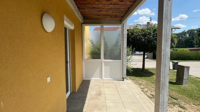 Überdachte Terrasse mit Holzbalken und Fliesenboden, angrenzend an die gelbe Hausfassade.