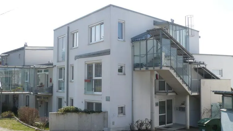 Modernes Mehrfamilienhaus mit weißer Fassade und verglasten Außentreppen in einer Wohnsiedlung.