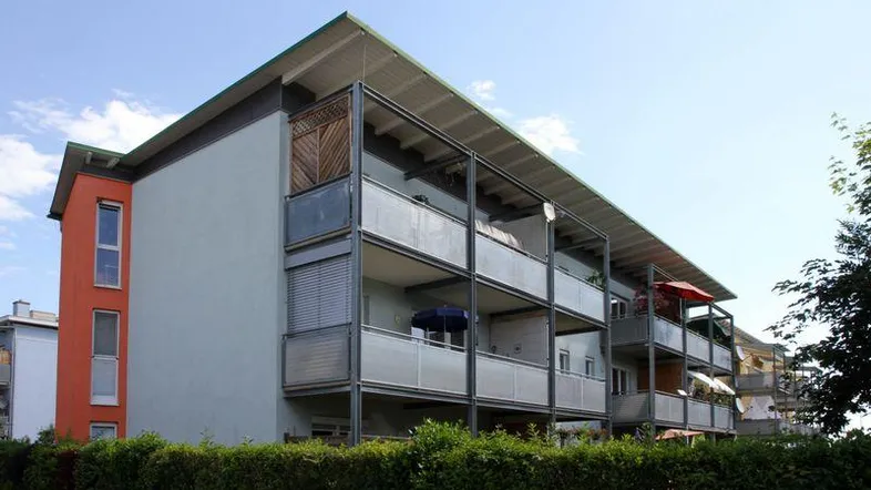 Modernes Mehrfamilienhaus mit Balkonen und einer gepflegten Hecke im Vordergrund unter blauem Himmel.