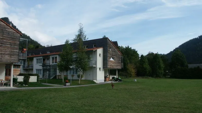 Modernes Wohngebäude mit Holzfassade, umgeben von einer weitläufigen Grünfläche und Bergen im Hintergrund.