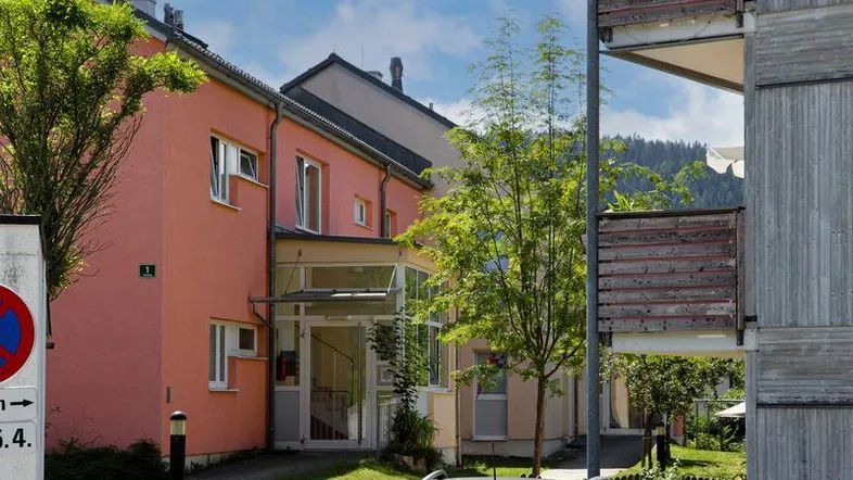 Mehrfamilienhaus mit lachsfarbener Fassade, umgeben von Grünflächen und Bäumen, mit Bergblick im Hintergrund.
