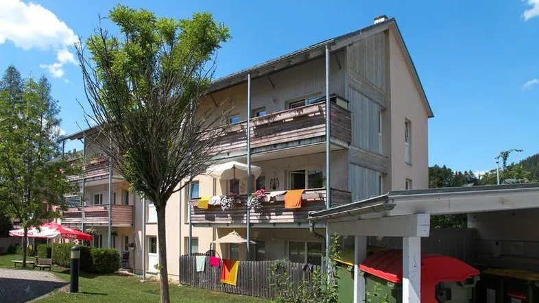 Mehrfamilienhaus mit mehreren Balkonen, umgeben von Grünflächen und Bäumen unter blauem Himmel.