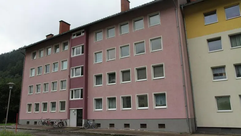 Mehrstöckiges Wohngebäude mit rosa und gelber Fassade, vielen Fenstern und Blick auf bewaldete Hügel.
