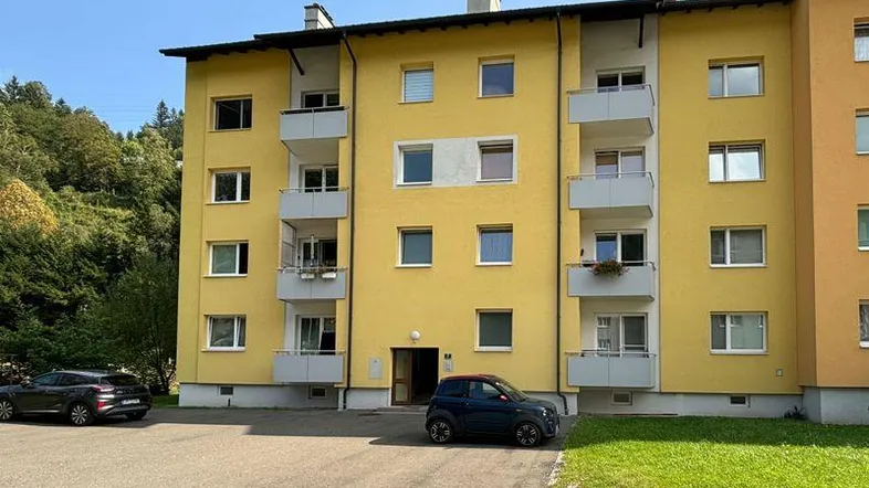 Mehrstöckiges Wohngebäude mit gelber Fassade, mehreren Balkonen und Parkplätzen davor, umgeben von grüner Natur.