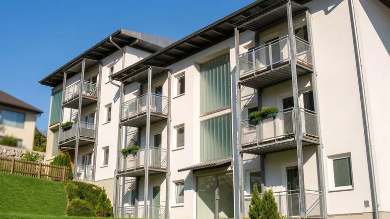 Modernes Mehrfamilienhaus mit weißer Fassade, mehreren Balkonen und grüner Umgebung.