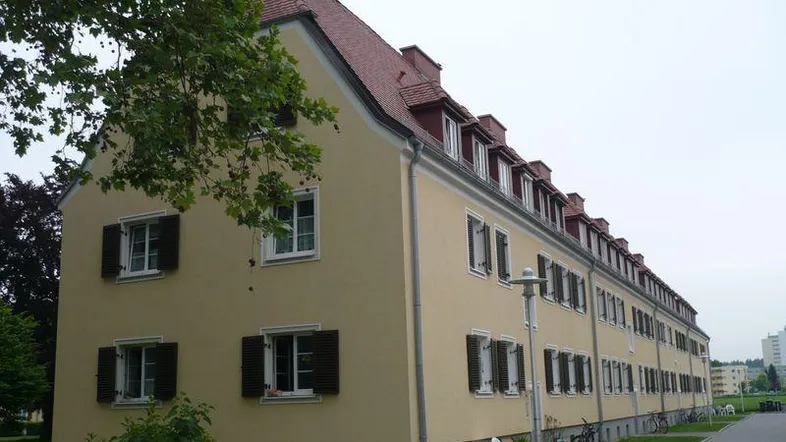 Langes Wohngebäude mit gelber Fassade, Satteldach und vielen Dachgauben, umgeben von Grünflächen.