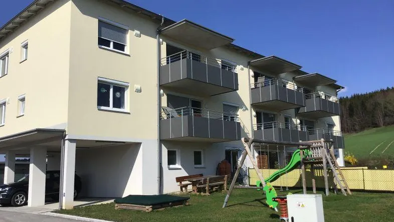 Modernes Mehrfamilienhaus mit Balkonen, Carport und Kinderspielplatz in ländlicher Umgebung.