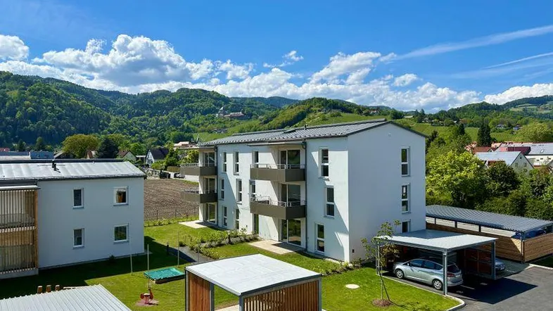 Modernes Mehrfamilienhaus mit Balkonen und Carports, umgeben von grüner Hügellandschaft unter blauem Himmel.