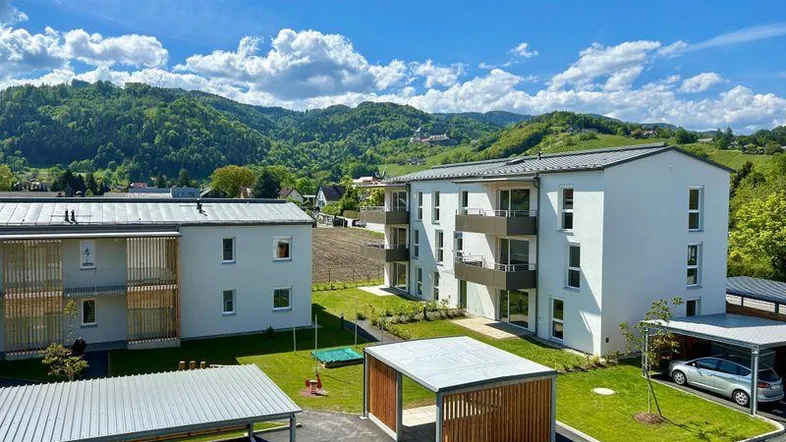 Moderne Wohngebäude mit Carports und Spielplatz, umgeben von grünen Hügeln und Bergen unter blauem Himmel.