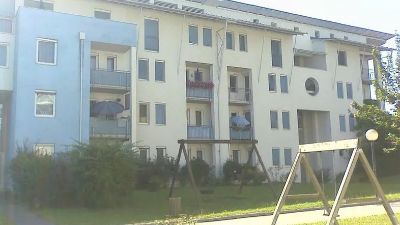 Mehrfamilienhaus mit blauer Fassadenakzentuierung, Balkonen und Kinderspielplatz im Vorgartenbereich.
