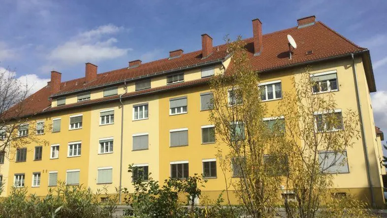 Mehrfamilienhaus mit gelber Fassade, rotem Ziegeldach und Rollläden an den Fenstern.
