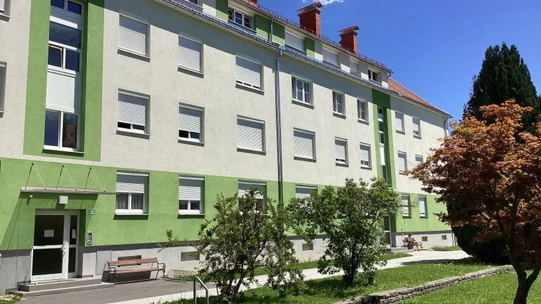 Mehrfamilienhaus mit grün-weißer Fassade und Rollläden, umgeben von einer kleinen Grünfläche.