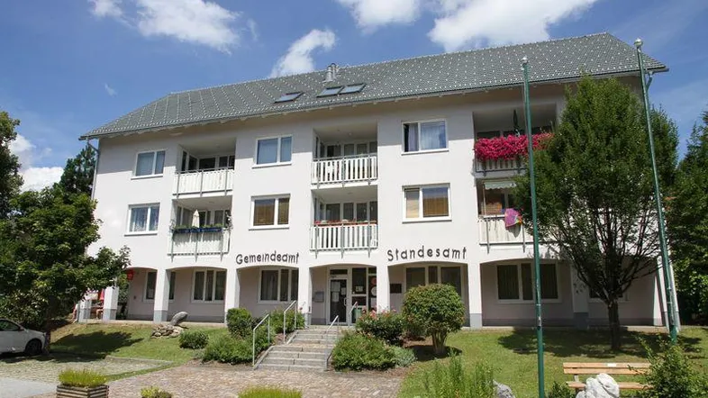 Mehrfamilienhaus mit weißer Fassade, Satteldach und Balkonen, umgeben von Grünflächen.