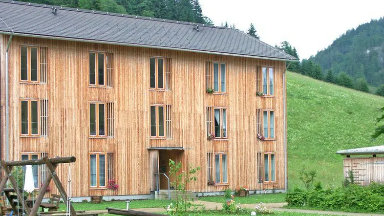 Modernes Holzhaus mit vielen Fenstern, umgeben von grünen Wiesen und Wäldern, mit einem Spielplatz im Vordergrund.