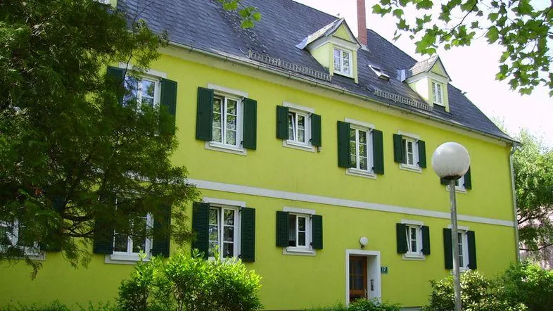Gelbes Wohngebäude mit grünen Fensterläden und Dachgauben, umgeben von Bäumen und Sträuchern.
