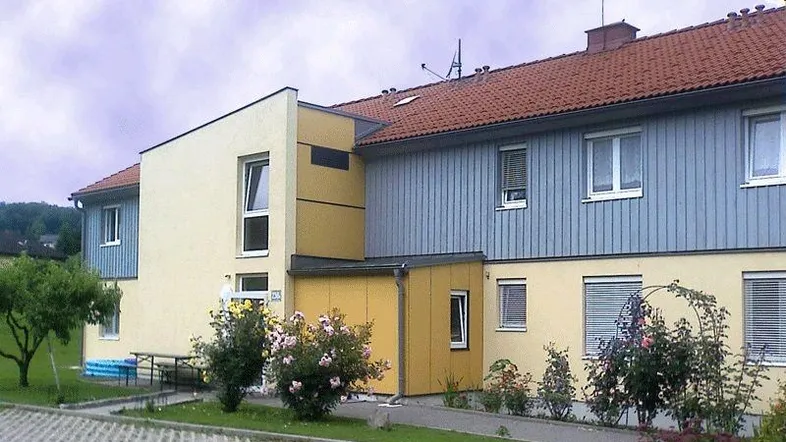 Mehrfamilienhaus mit gelber und blauer Fassade, umgeben von Grünflächen und Büschen, mit barrierefreiem Zugang.