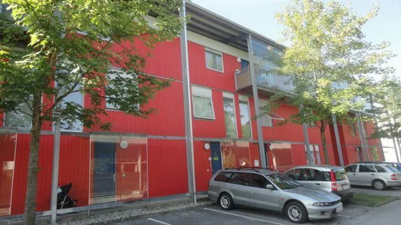 Modernes Wohngebäude mit leuchtend roter Fassade, umgeben von Bäumen und Parkplätzen.