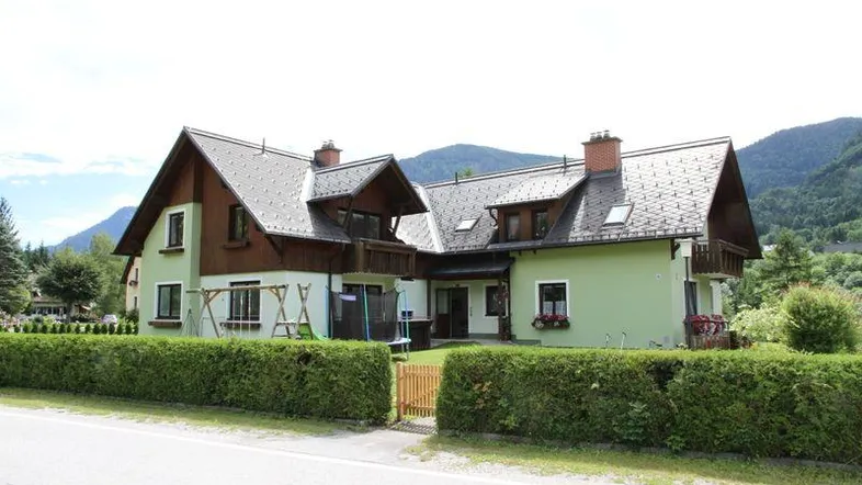 Grünes Einfamilienhaus mit Satteldach, umgeben von Hecken und Bergen im Hintergrund.