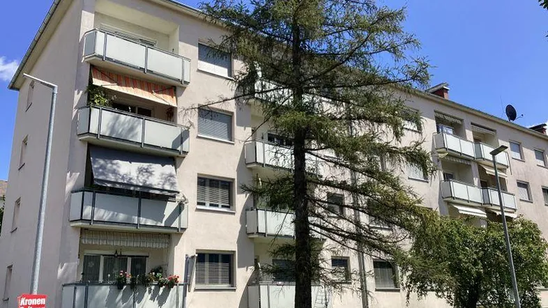 Mehrfamilienhaus mit mehreren Balkonen und grüner Umgebung unter blauem Himmel.