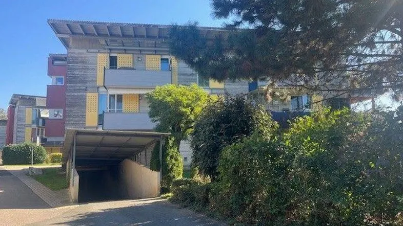 Mehrfamilienhaus mit Carport und begrüntem Vorgarten unter blauem Himmel und Sonnenschein.