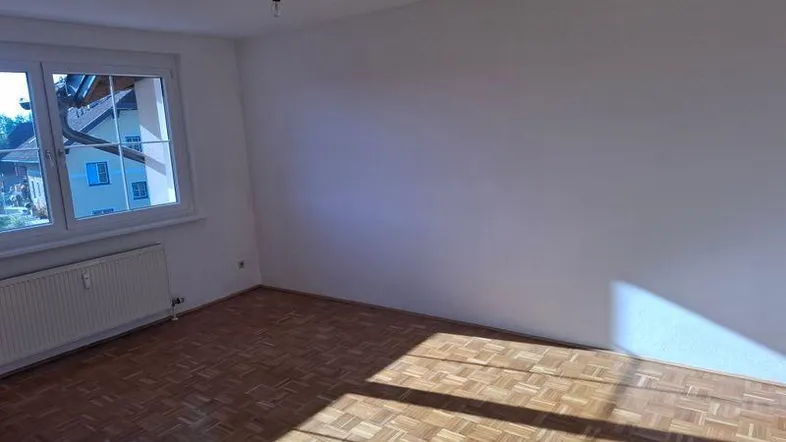 Helles, leeres Zimmer mit Parkettboden und großem Fenster, das viel Tageslicht hereinlässt.