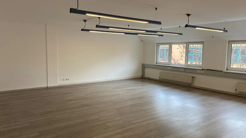 Geräumiger, leerer Büroraum mit modernen Hängelampen, großen Fenstern und hellem Holzboden.
