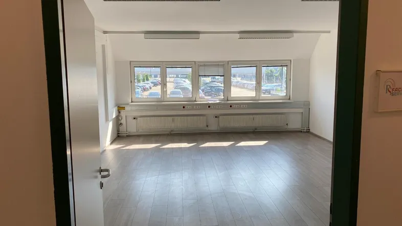 Geräumiges, leeres Büro mit großen Fenstern, die viel Tageslicht hereinlassen und einen Blick auf den Parkplatz bieten.