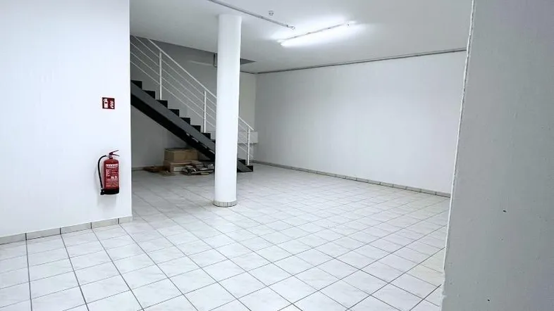 Großer, leerer Raum mit weißen Fliesen, einer Säule und einer Treppe im Hintergrund, bereit zur Nutzung.