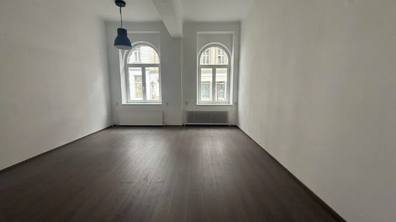 Heller, leerer Raum mit dunklem Holzboden, weißen Wänden und zwei großen Bogenfenstern.